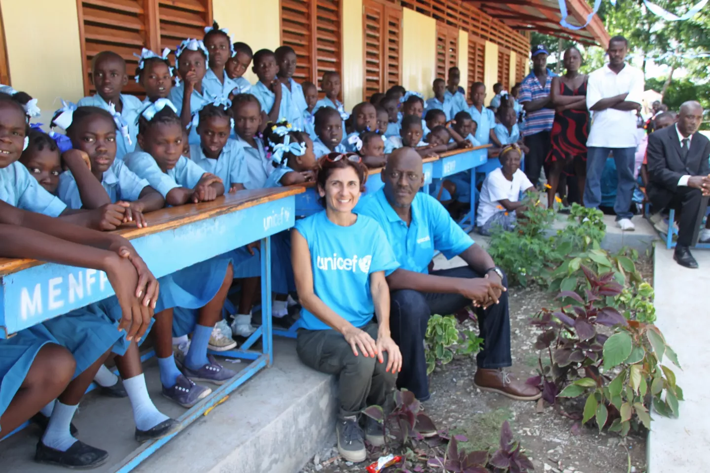 A woman and  man, both wearing t-shirts that read "UNICEF", sitting in front of a group of children. They are all smiling.