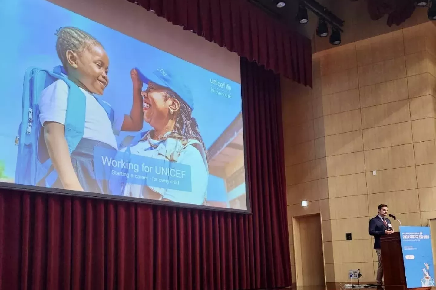 A man on a podium speaking. In the background a photo of a woman holding a child . Overlay text reads: "Working for UNICEF. Starting a career, for every child"