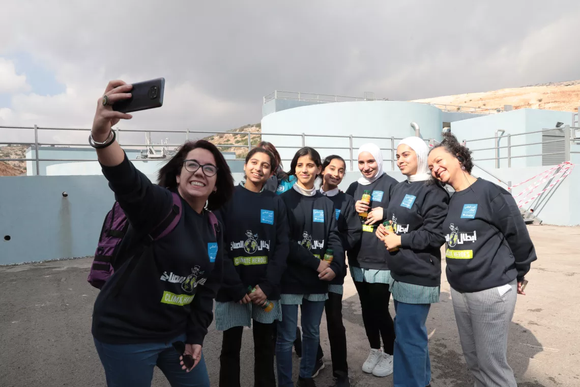 A group of young women stand together and smile while one of them holding a mobile phone takes  a selfie photo