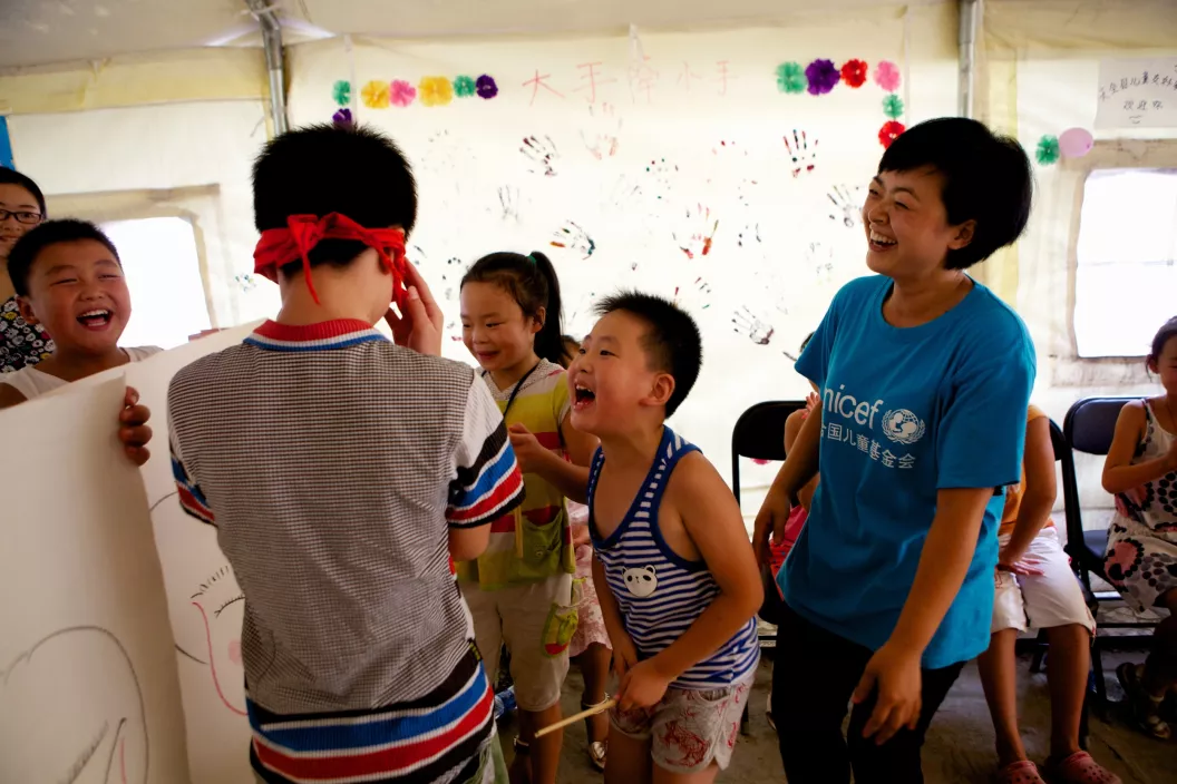 a child blindfolded plays with other children and a woman wearing a UNICEF t-shirt is looking at them laughing