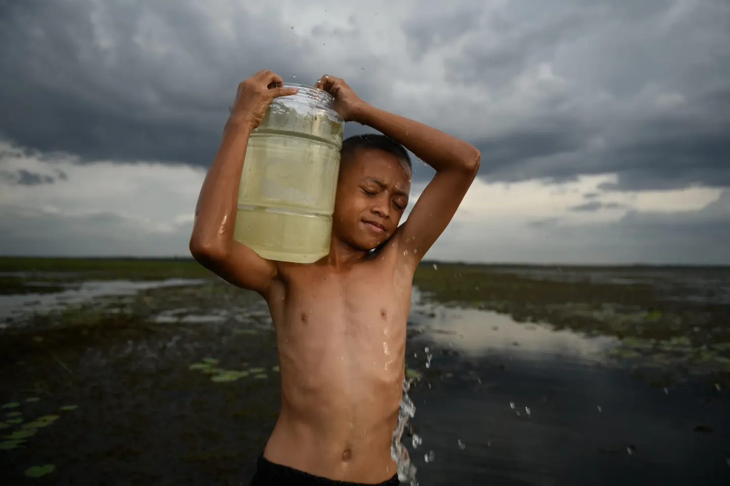 Nattapong Saichumdee (Bank) stands in a shallow, flooded field, holding a large container of water over his shoulder.