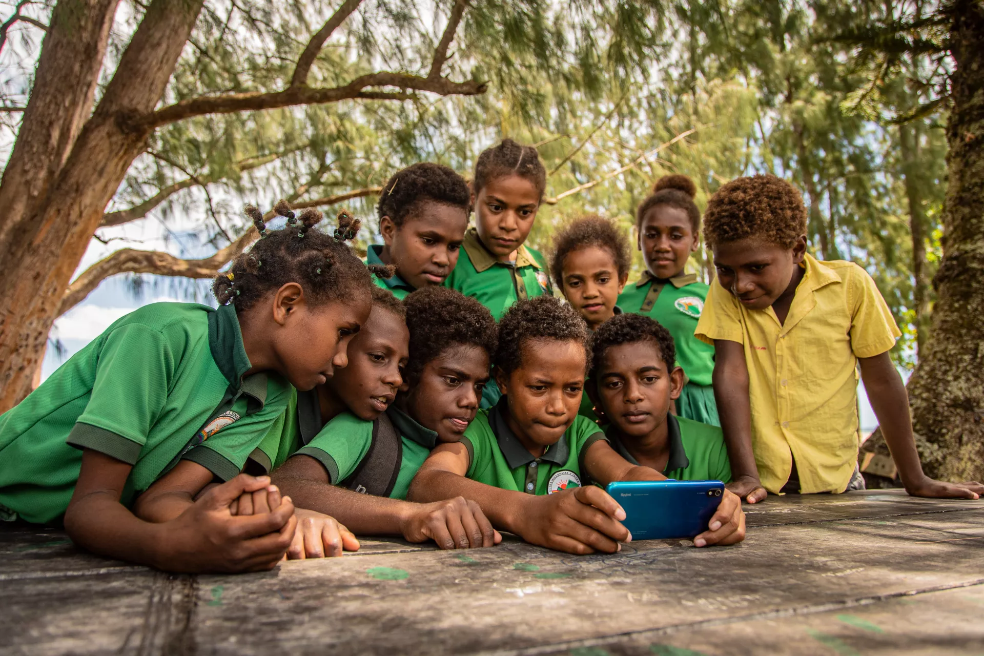 Boys and girls gather around to watching one of them playing a video game on his mobile device.