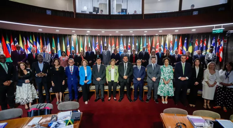 Hombres y mujeres de pie en una sala de reuniones, delante de banderas de Am&eacute;rica Latina y el Caribe