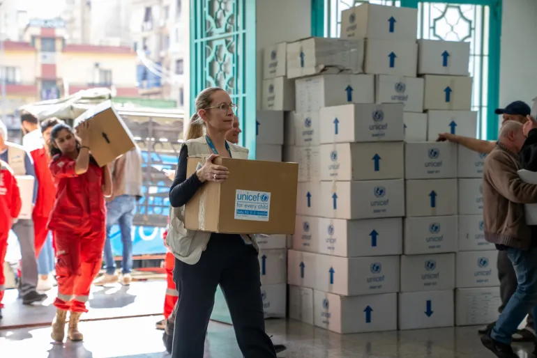 Representante Adjunta de UNICEF L&iacute;bano, carga una caja de suministros de emergencia con la etiqueta 'Baby Kit, 0-3 months' en una escuela p&uacute;blica del Monte L&iacute;bano habilitada como refugio. Al fondo, personal humanitario apila cajas de UNICEF.