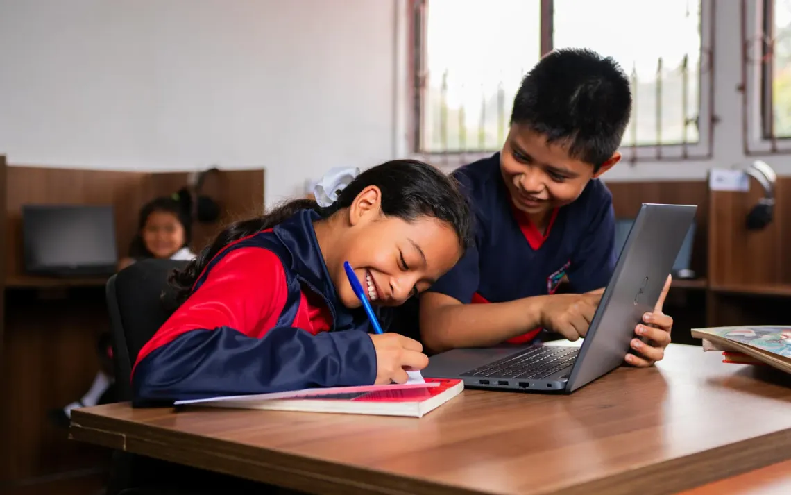 Two children smile, while learning together in a classroom using notebook, pen and a laptop