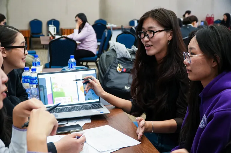 Three girls lean in toward a laptop showing the Oky app while one young women gestures with a pen, mid-discussion with teammates at a worktable at the Oky Mongolia Hackathon.