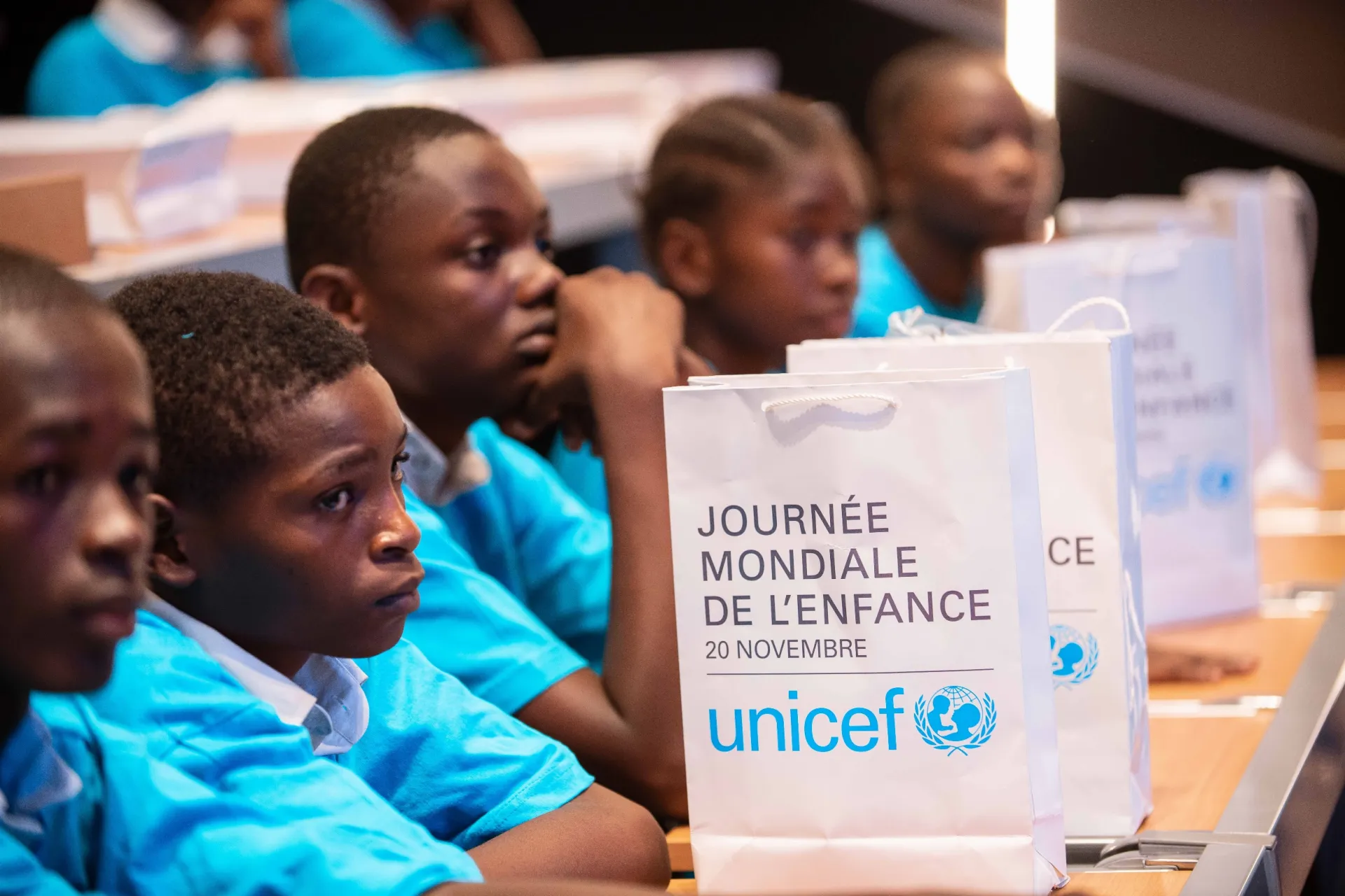 Un sac portant l'inscription Journ&eacute;e mondiale de l'enfance pos&eacute; sur un banc derri&egrave;re lequel sont assis des enfants