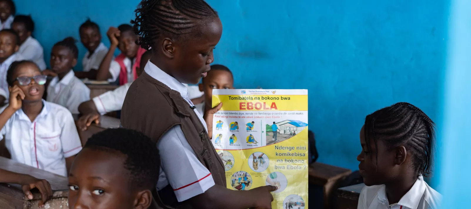 Une fille tient une affiche Ebola et la montre &agrave; une &eacute;l&egrave;ve assise sur un banc