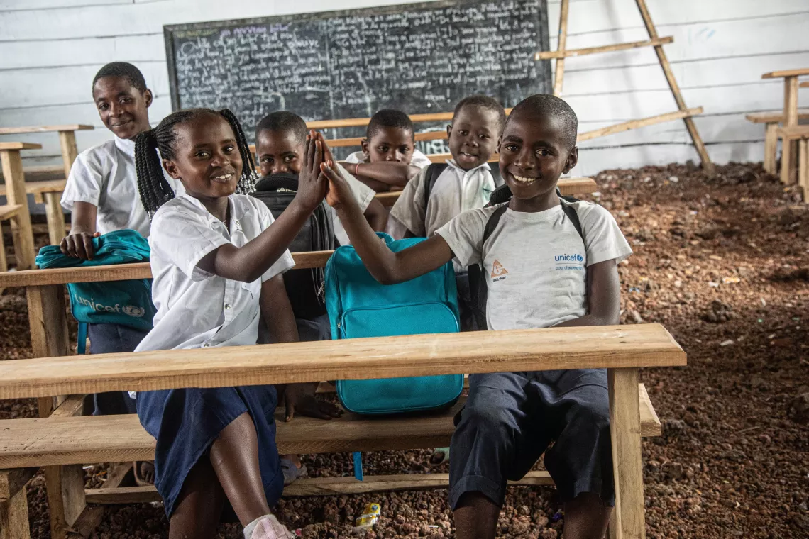 Two children high-five while sitting on a wooden bench in a temporary classroom