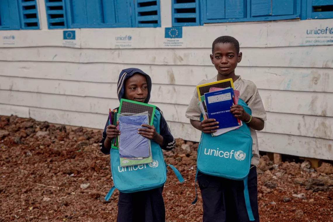 Two children hold their new school supplies in front of a temporary classroom.