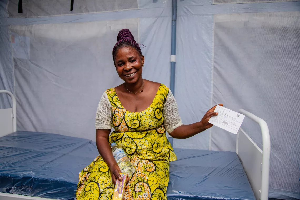 Une femme, assise sur un lit, tient un certificat de vaccination en souriant