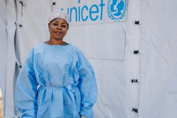 Une professionnelle de santé pose devant une tente UNICEF, vêtue d'une blouse de protection bleue.