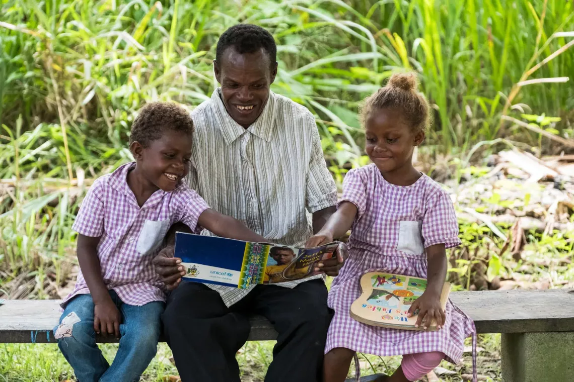A father reading a book to his 6 year old twin children Chris (left) and Florence (right) near Vutu Primary School in Guadalcanal Province, Solomon Islands.
