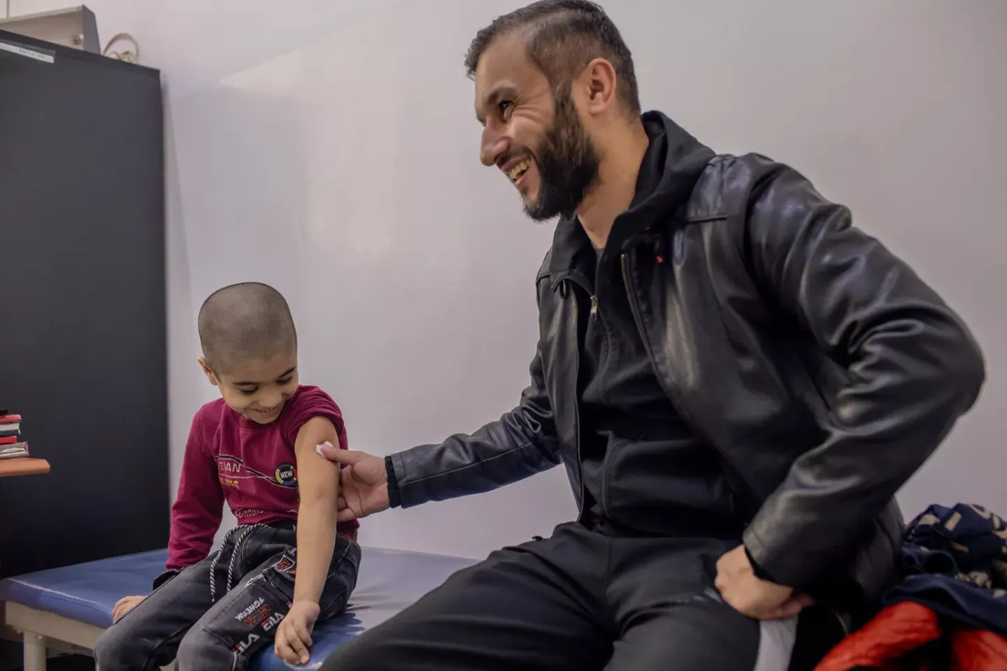 Six-year old Muso Ahliddinzoda smiles as he looks down at his arm, held by his father, after  the boy has his routine vaccinations at a health centre in Dushanbe, Tajikistan. March 2022.