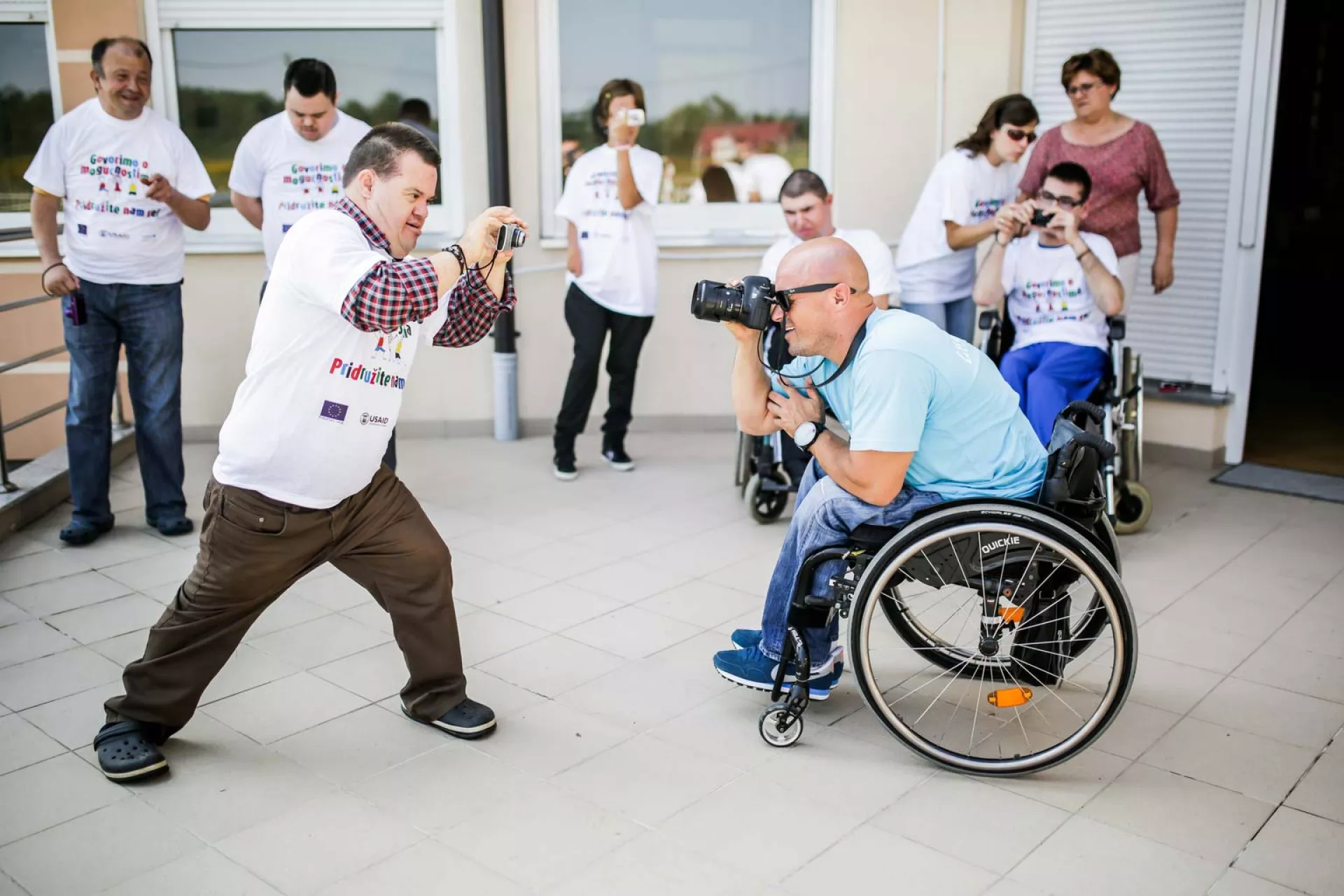 Two young men with disabilities playfully point cameras at each other.