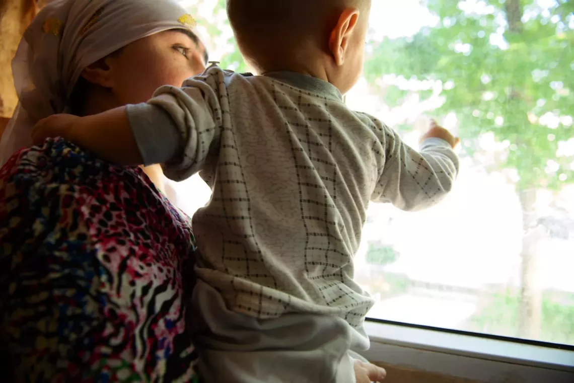 Mother Maqsuda*, 30, holds her 1-year-old son Jovaid as&nbsp;she looks out from a window in her home in Dushanbe, Tajikistan. December 2014. 