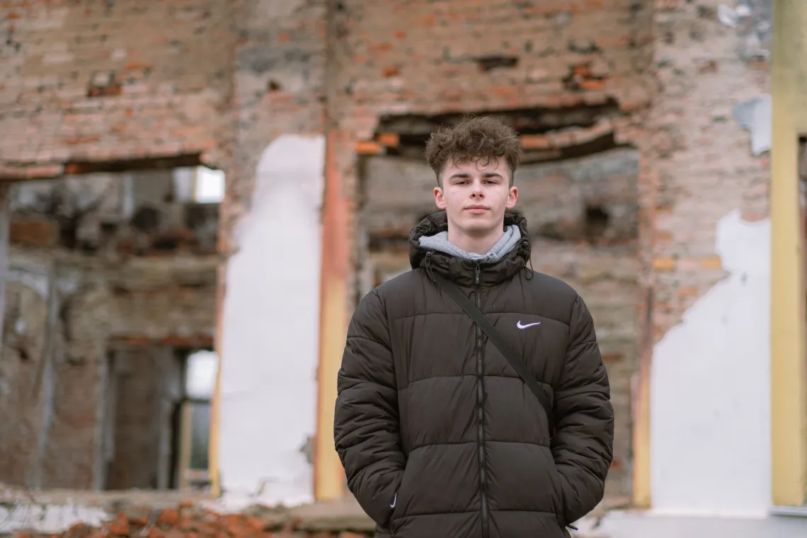 Yaroslav (16) stands against the background of a destroyed school, he used to study in.