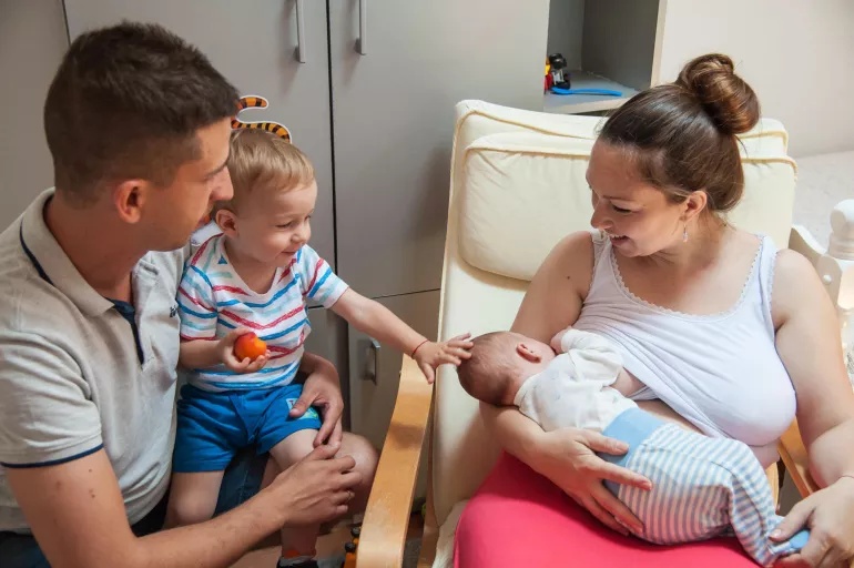Mother breastfeeds her baby, while the father and the older son support them.