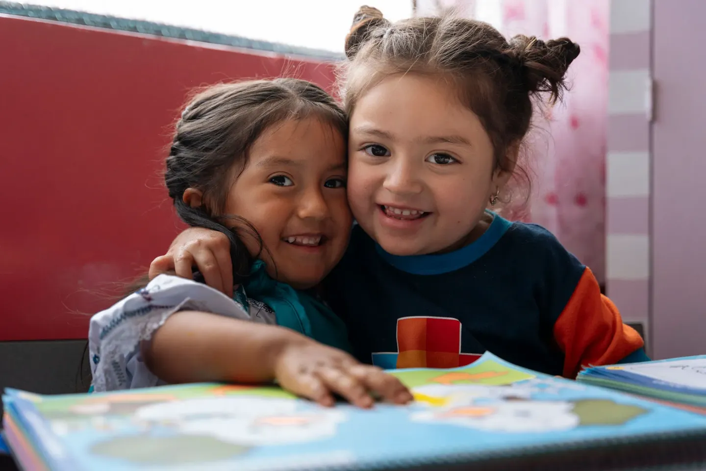 Dos ni&ntilde;as sonrientes se abrazan junto a una mesa con dibujos y crayones en un aula colorida.