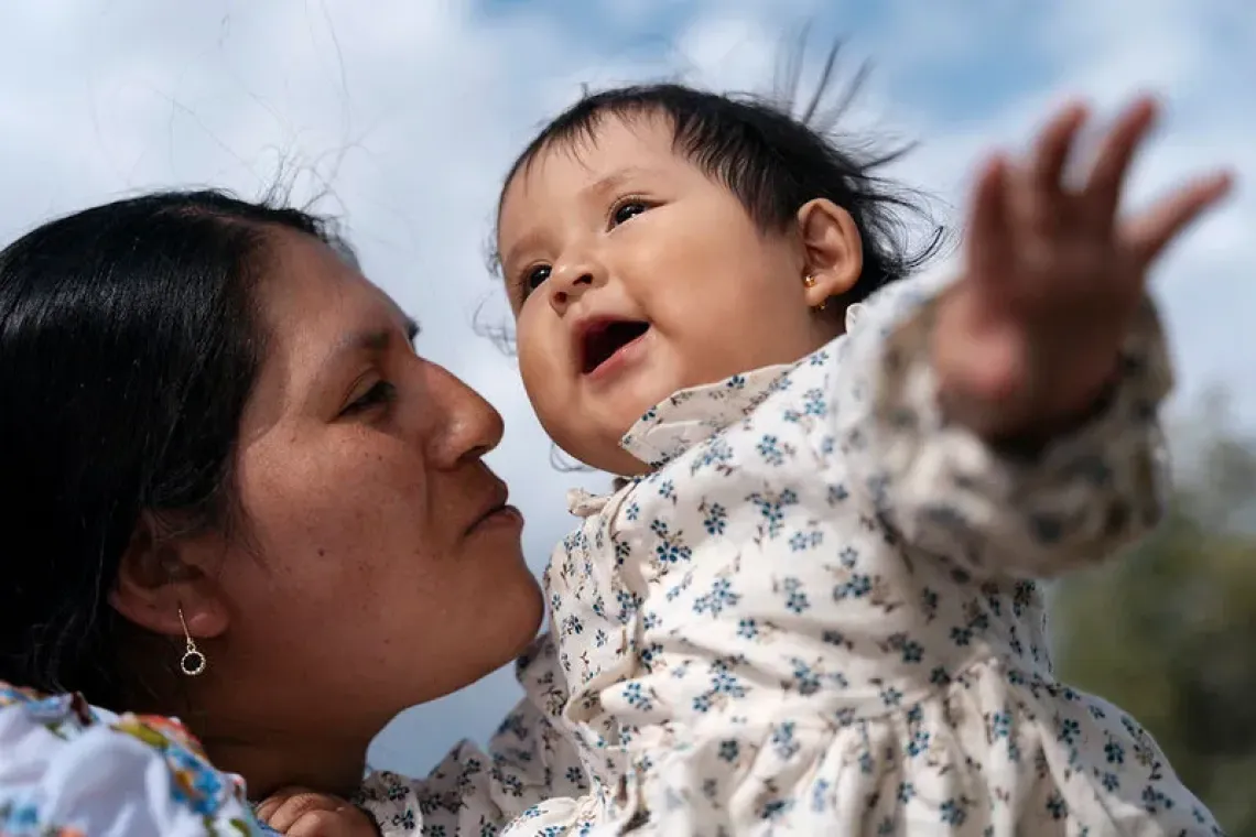 Mujer levanta y besa a un beb&eacute; sonriente bajo un cielo azul.