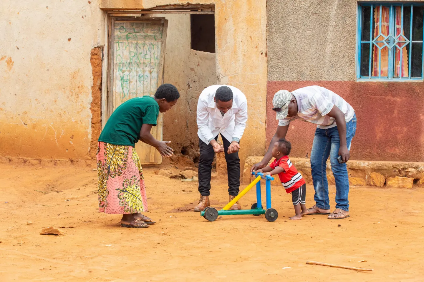 Caregivers encourage a child to stand and push a tricycle meant to help accelerate walking.