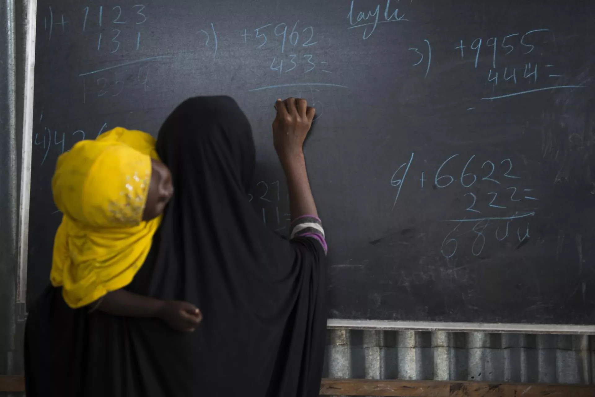 Ismahan, 13, holding her sleeping 3-year-old sister, works on an arithmetic problem at the blackboard, Somalia.
