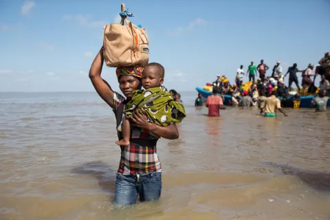 A mother carries supplies and her child as she wades through water after disembarking from a boat