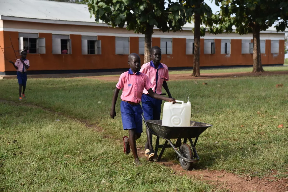 Two students push a wheelbarrow holding a jerry can of water