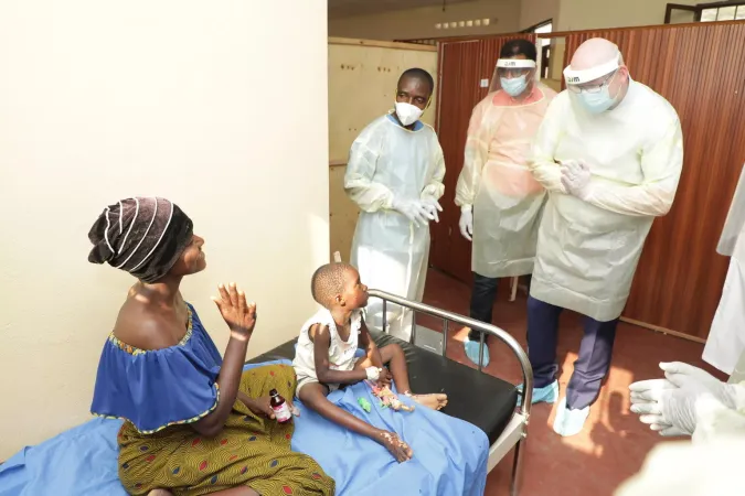 A mother and her child sit on a hospital bed speaking to healthcare workers.