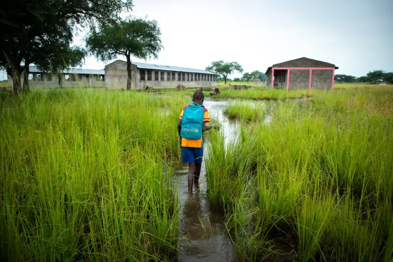 A child wearing a UNICEF blue backpack walks towards a school amidst a flooded field