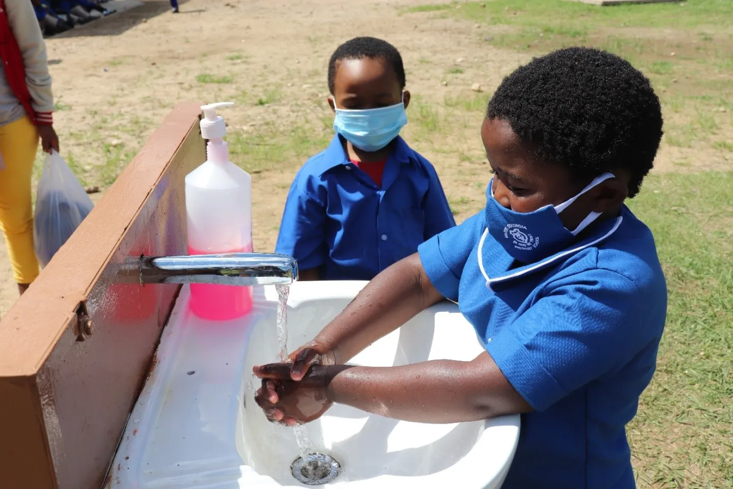 Two children outdoors, one washing hands with running water
