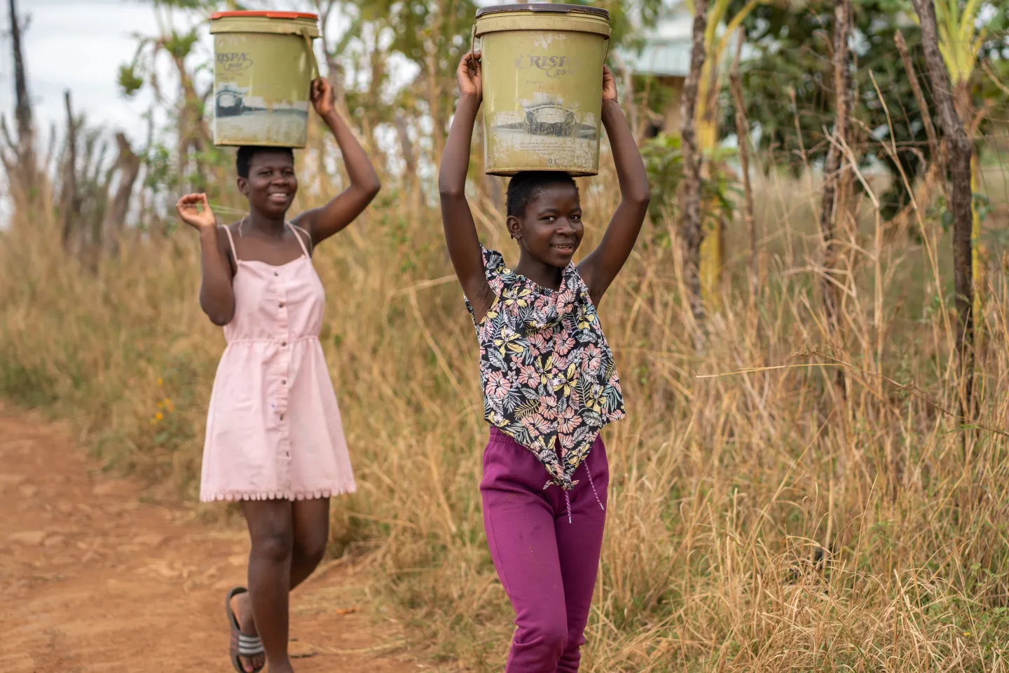 Two girls carrying water buckets and walking along a dusty road.