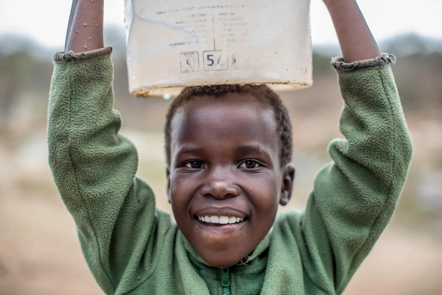 A young child smiles in this close-up image, holding a white bucket on their head