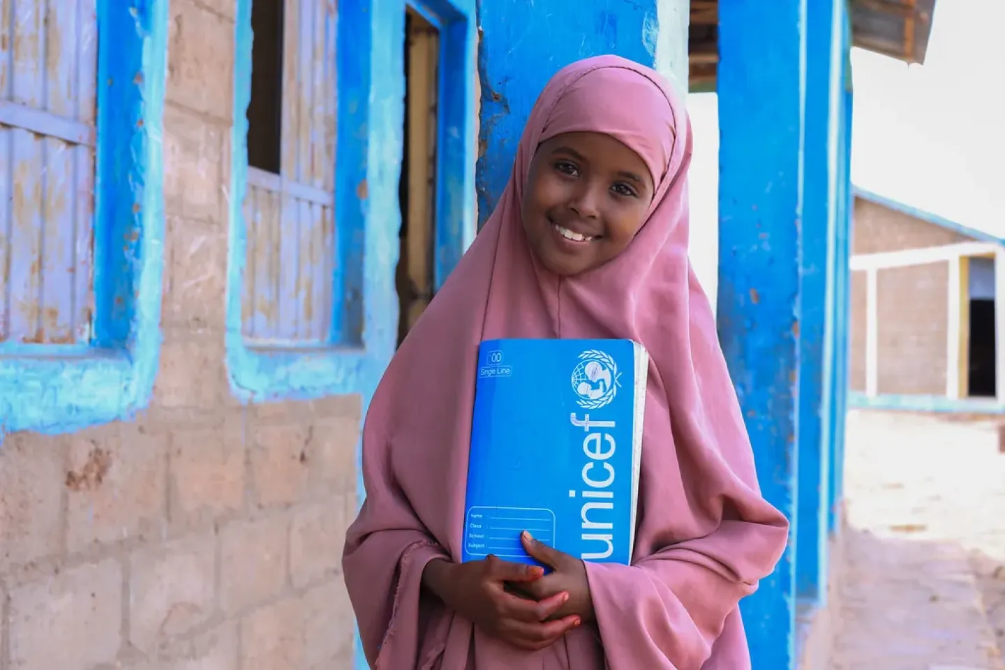 A little girl holding her note book close to her heart and posing for the camera outside of her classroom