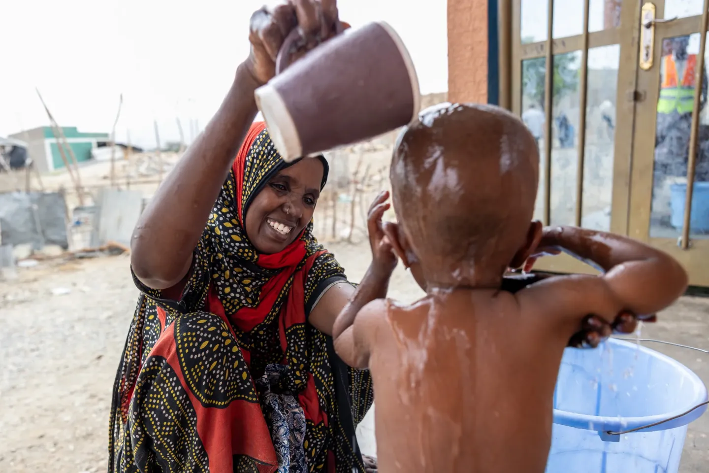 Smiling caregiver pours water over a young child during bath time outdoors.