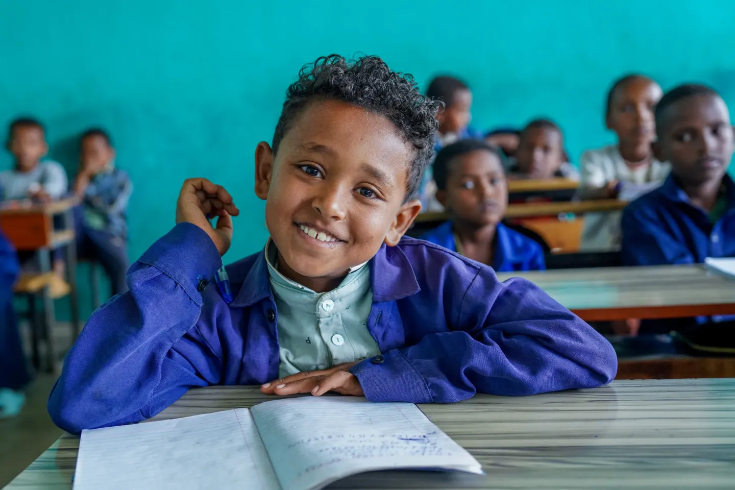 A little boy sitting on his desk and smiling in his classroom