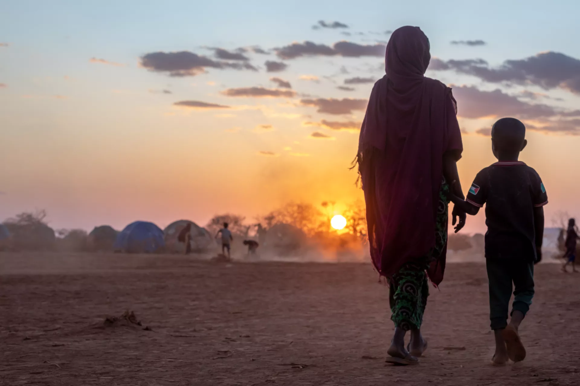 A mother and a child walk together in Bul'ad IDP camp as the sun sets in the horizon. 