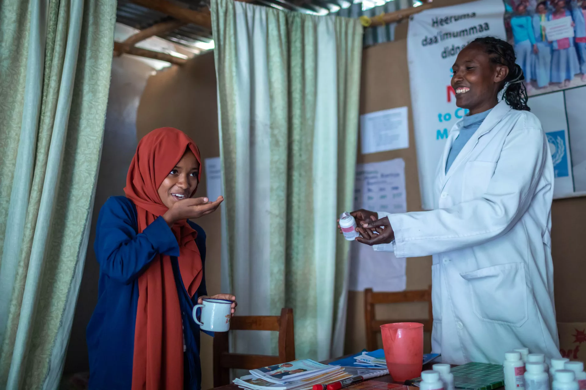 A girl with a dark orange head scarf and navy dress holds a mug in her left hand, while her right hand hovers near her mouth. Beside her, a woman in a white coat holds tablet container. 