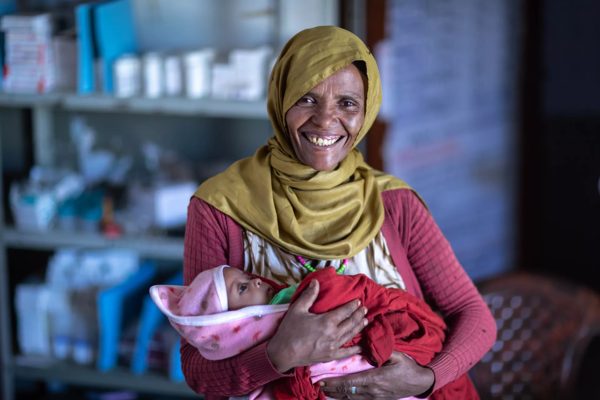 A woman with a yellow head scarf smiles widely as she holds her sleeping baby. Behind her are bottles of medication sitting on a shelf.