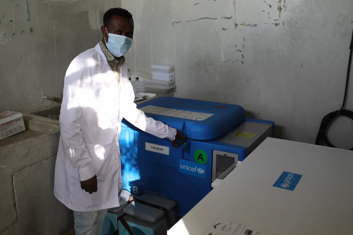 A male health worker wearing a white coat gestures towards a large blue refrigerator.