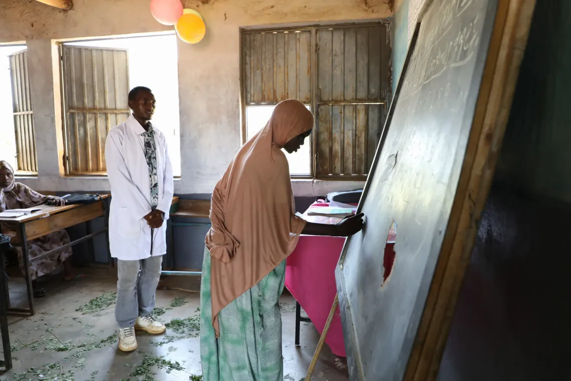 Rawiya writing on her classroom blackboard, while her teaching, Abdelnur Abdellah watches on.