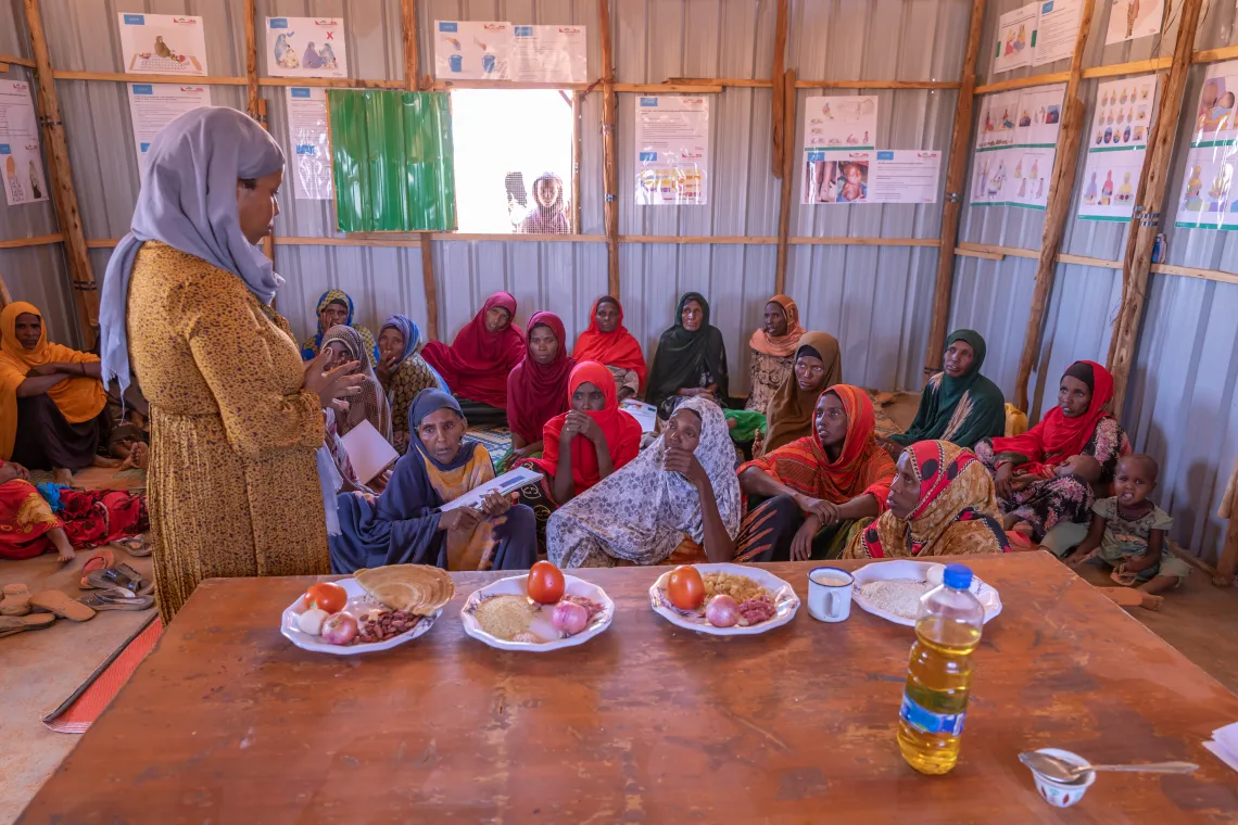 A trainer explain how to prepare nutritious food for a group of women sitting in a room while food items are displayed on a table