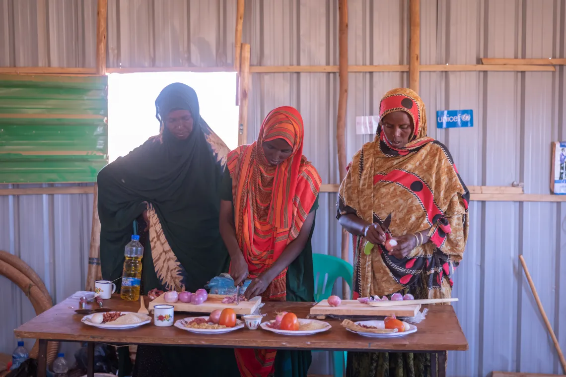Three women preparing food; chopping onions on a board. Plates with food items are displayed in front of them.  