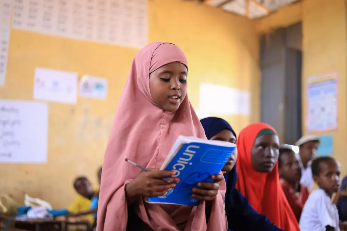 A young girl in her classroom, standing at her desk and reading from her exercise book. Her classmates sit behind her in the background.