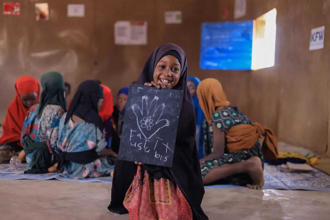 A girl child holding her drawing on a chalk board in a SPEED school classroom. Her classmates in the background.