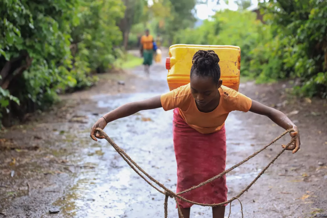 A young girl carrying a jerry can on her back and making a rope to strap it. 