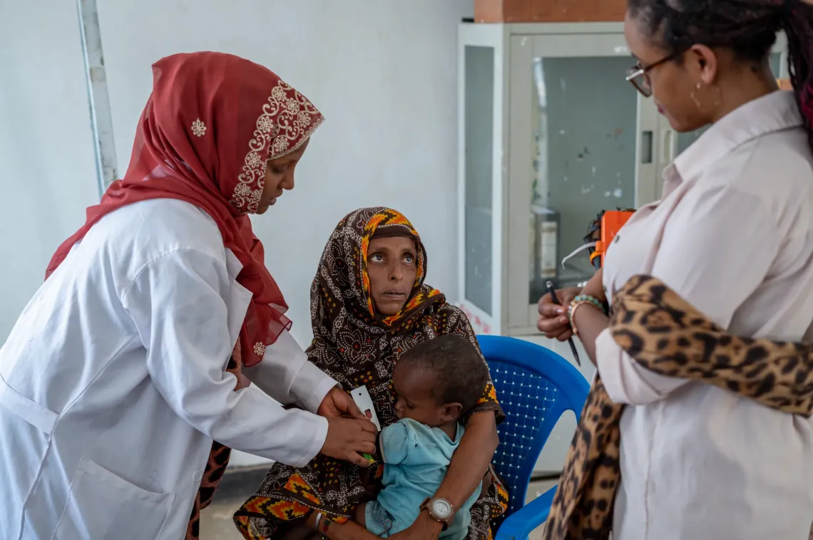 A mother sitting and holding her baby boy, while a health worker is taking a MUAC measurement 