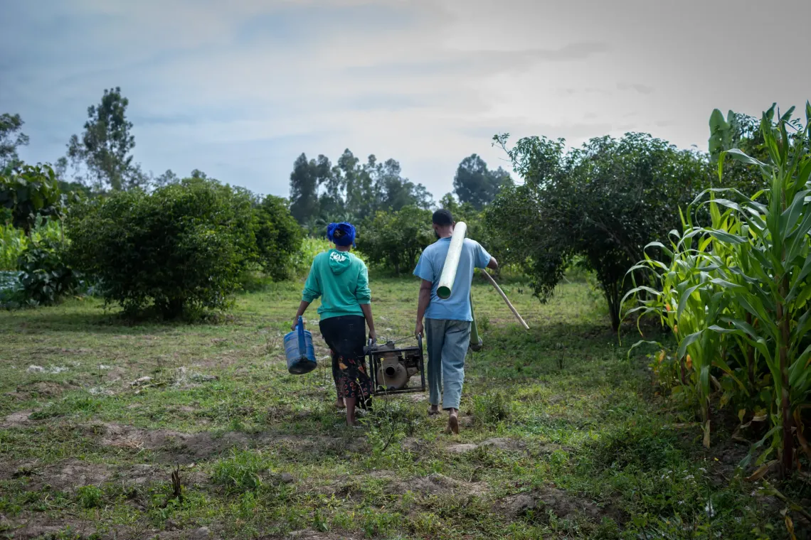 A husband and wife walk towards their farm carrying a water pump