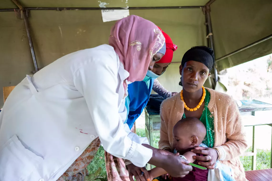 A mother holding her son while a health worker monitors his weight using MUAC measurement. 