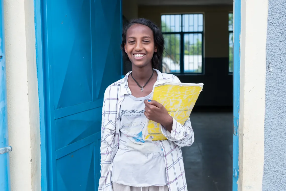A young girls smiles holding her books by the door of her class room